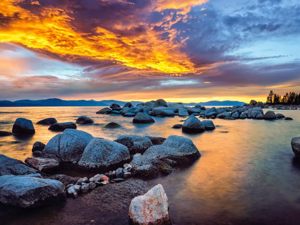 Lake Tahoe, Nevada y California, EE.UU., tomadas en Zephyr Cove en una espectacular puesta de sol con aguas tranquilas que rodean grandes rocas y un cielo amarillo.