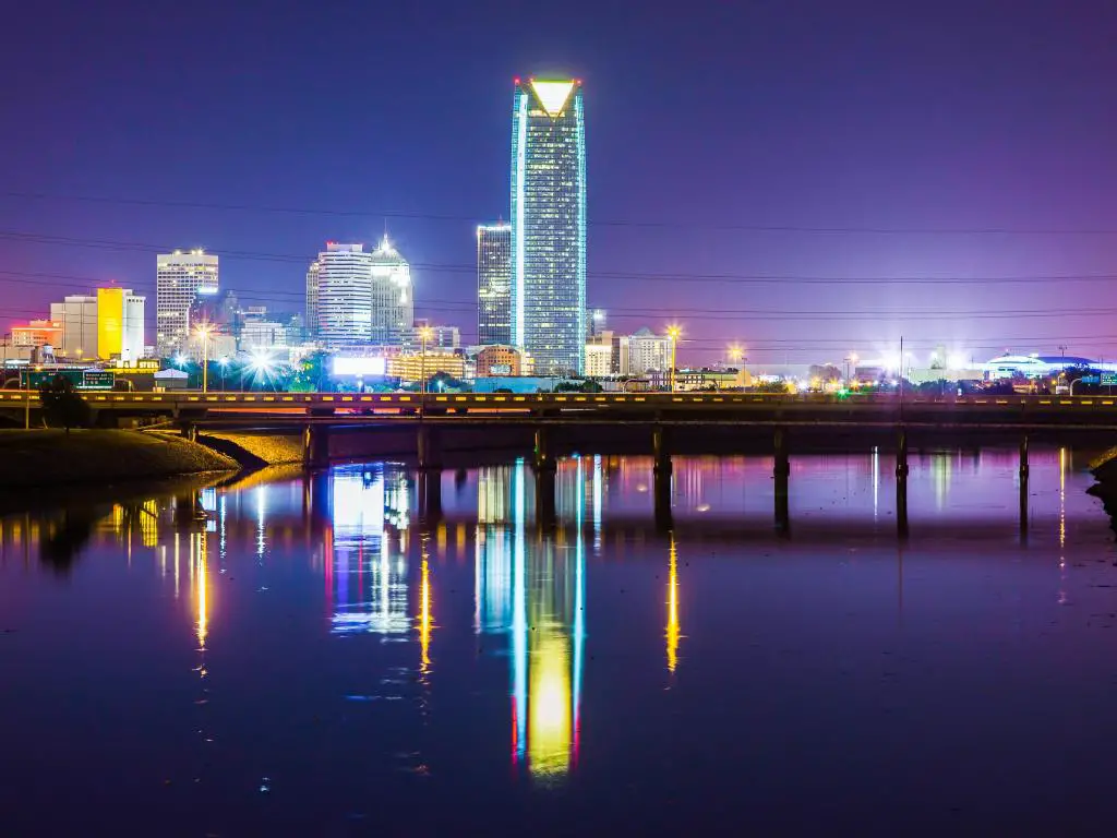 Oklahoma City, EE. UU. Con el horizonte nocturno reflejado en el agua debajo, un puente que cruza entre la ciudad y el cielo con un tono púrpura y azul. 