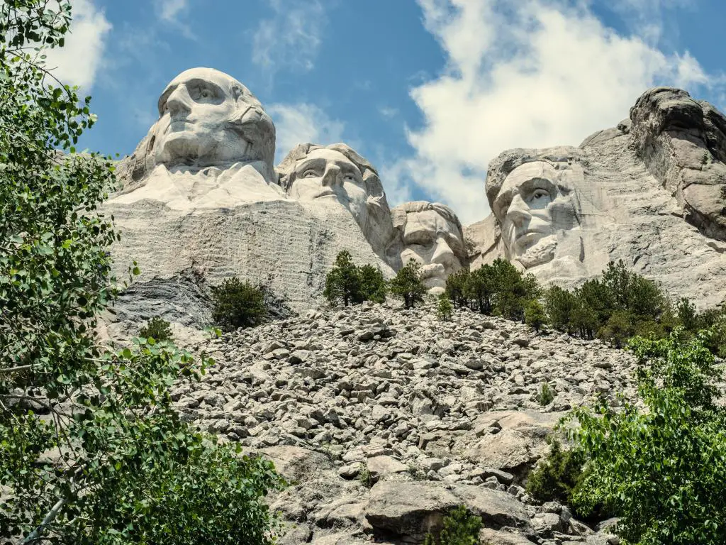 Monte Rushmore, Dakota del Sur, EE. UU. con los famosos presidentes tallados en Black Hills, árboles en primer plano y un cielo nublado pero soleado arriba.
