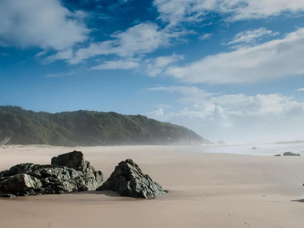 Amplia playa de arena dorada con una sola roca en primer plano y un promontorio cubierto de árboles a lo largo del horizonte