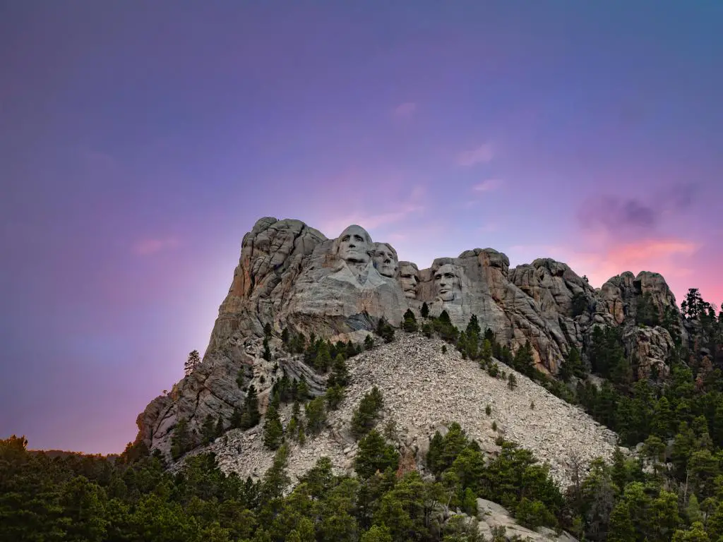 Monte Rushmore, EE. UU. con las Black Hills ajardinadas del Monte Rushmore contra un cielo crepuscular de color rosa y púrpura y árboles en primer plano.