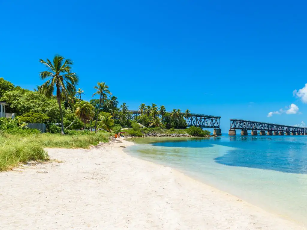 Parque Estatal Bahia Honda, Big Pine Key, Florida con una costa tropical y una playa en primer plano y un puente alto en el fondo bajo un cielo azul.