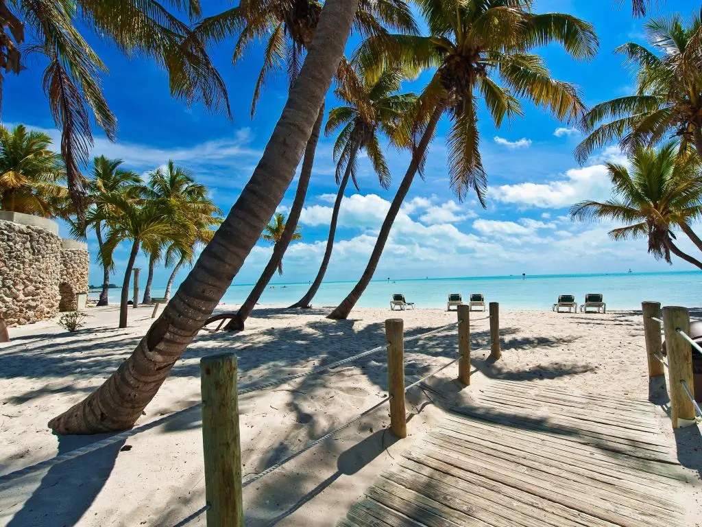 Key West, Florida, con un puente peatonal en primer plano que conduce a la playa con palmeras a ambos lados y el agua turquesa a lo lejos en un día soleado.