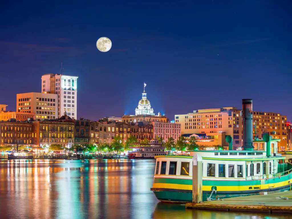 Edificios históricos frente al mar vistos al otro lado del río por la noche con un barco fluvial en primer plano y la luna