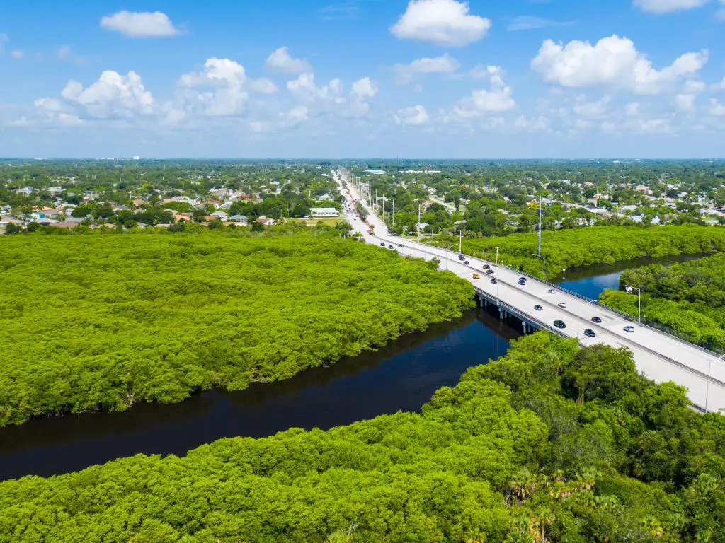 Port St. Lucie, Florida, con una densa vegetación en primer plano, un puente de carretera sobre un río y más vegetación en la distancia en un día soleado. 