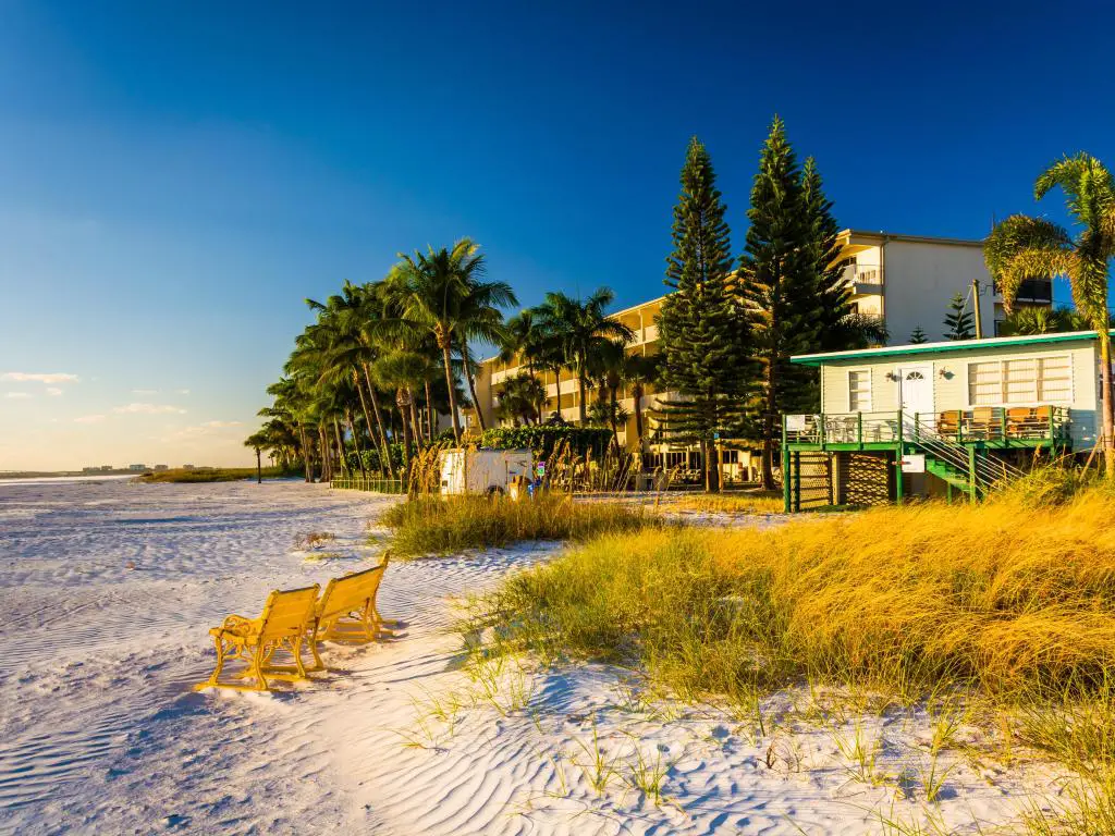 Fort Myers Beach, Florida, con dunas de arena, sillas frente al océano y edificios de playa detrás de una hilera de palmeras. 