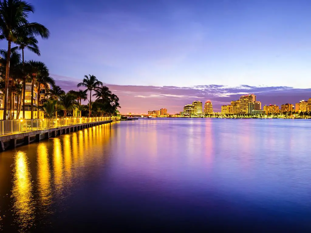 West Palm Beach, Florida, por la noche con las luces de la ciudad reflejándose en el agua en primer plano y altas palmeras que bordean el borde del agua.