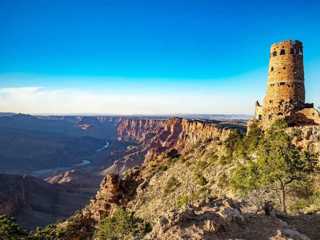 Grand Canyon Village, EE. UU. Con el Gran Cañón a lo lejos y una antigua torre de vigilancia en ruinas en primer plano sobre un cielo azul.
