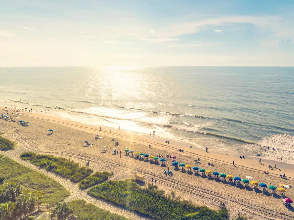 Vista aérea de Myrtle Beach, Carolina del Sur al atardecer con coloridas sombrillas en la playa, gente y vegetación en primer plano.