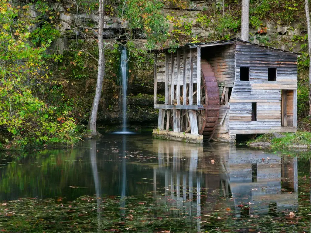 Antiguo molino de agua de madera con vegetación verde y una pequeña cascada, reflejada en el lago tranquilo