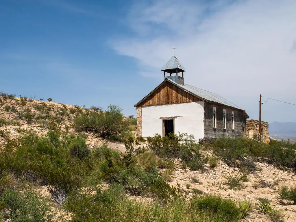 Pequeña iglesia blanca en la ladera de tierra seca con arbustos y cielo azul nublado