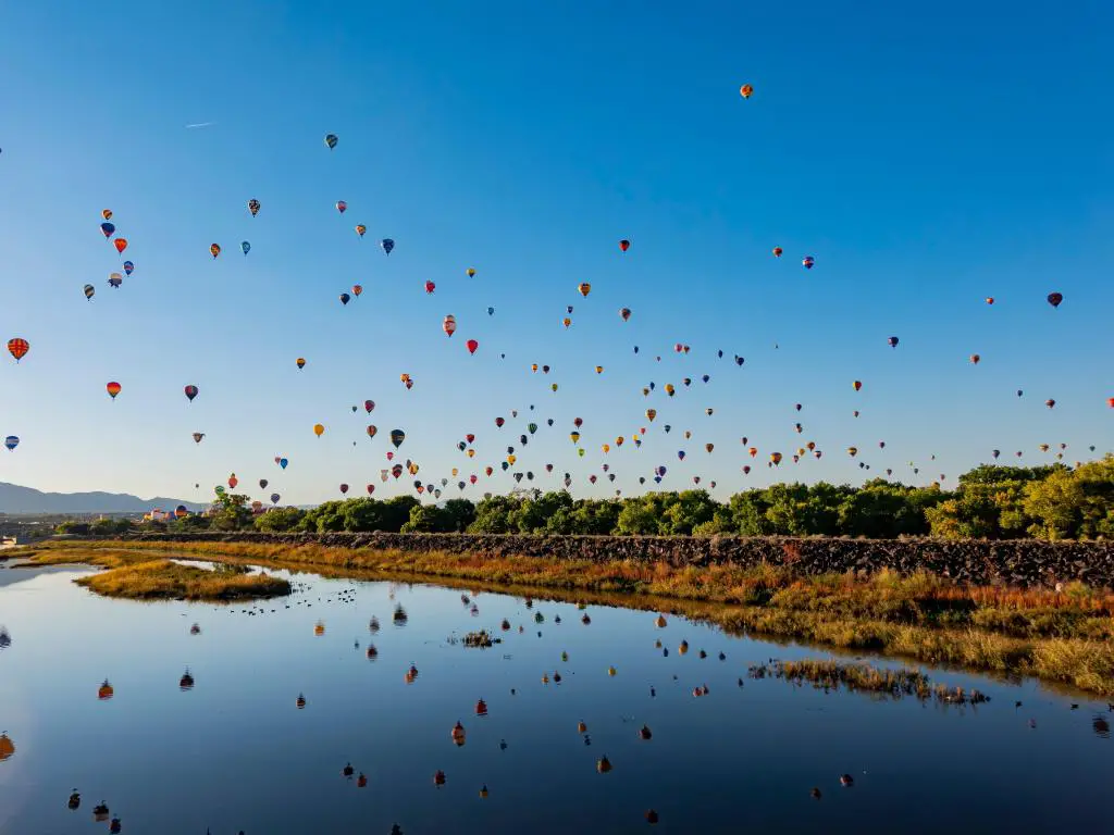 Muchos globos de aire caliente de colores brillantes vuelan a través del cielo azul sobre un lago, reflejado en el agua debajo