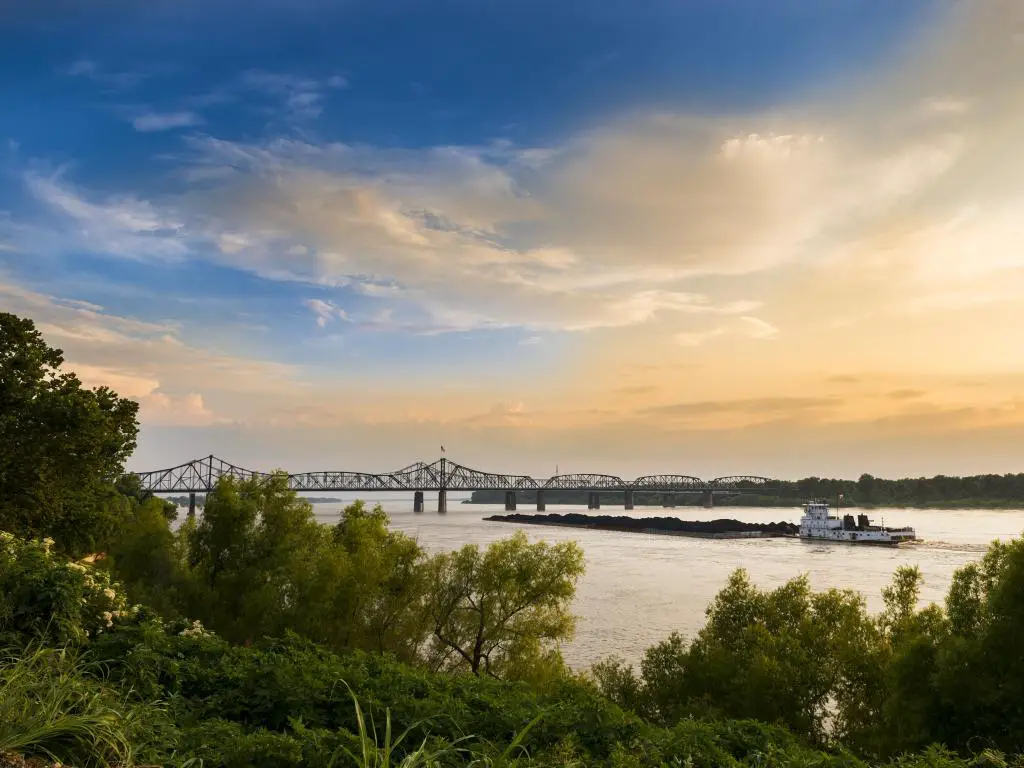 Puente de Vicksburg en Vicksburg, Mississippi con un barco en el río Mississippi, follaje verde en primer plano y el puente en la distancia al atardecer.