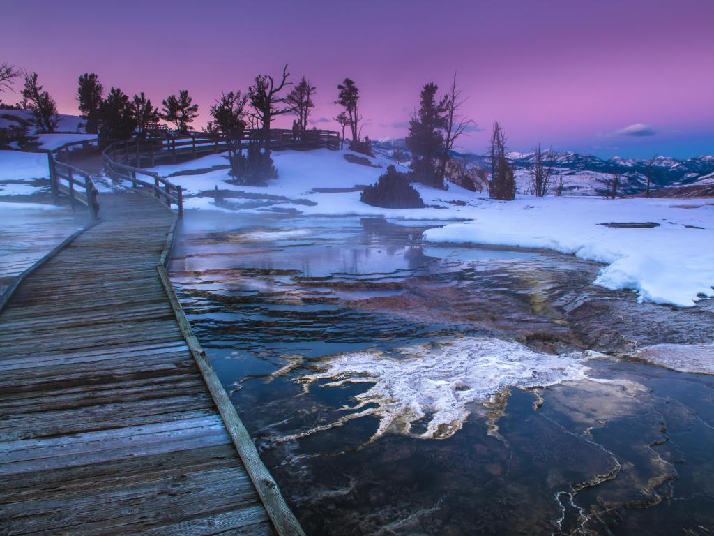 Camino que pasa por agua parcialmente congelada con montañas nevadas en la distancia y una puesta de sol rosa