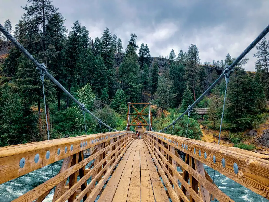 Dos personas al otro lado del puente colgante de madera con altos árboles verdes en la espalda y agua embravecida en Riverside State Park, Spokane, Washington