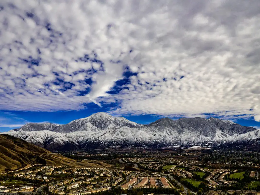 Una impresionante vista aérea de las montañas San Gorgonio y Little San Bernardino cubiertas de nieve en un día de invierno con cielos azules de nubes blancas y grises.