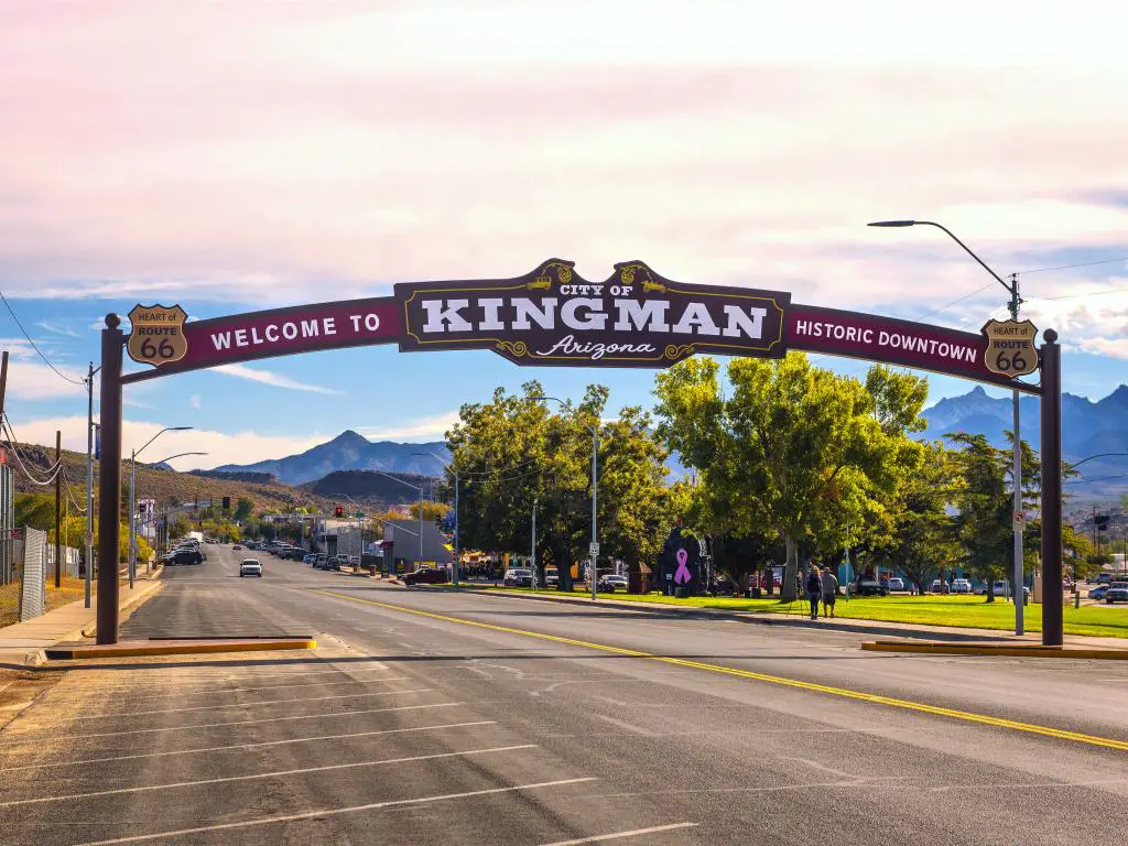 Un letrero de arcos ubicado en la ruta histórica 66 Kingman, Arizona, con autos conduciendo por la carretera, gente caminando en la acera bajo la sombra de los árboles y la vista de la montaña desde la distancia