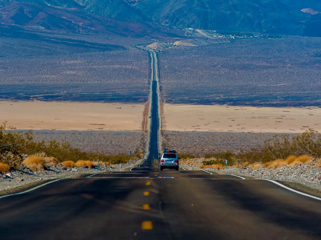 Carretera recta del Parque Nacional del Valle de la Muerte con vistas a las montañas y algunas hierbas a ambos lados.
