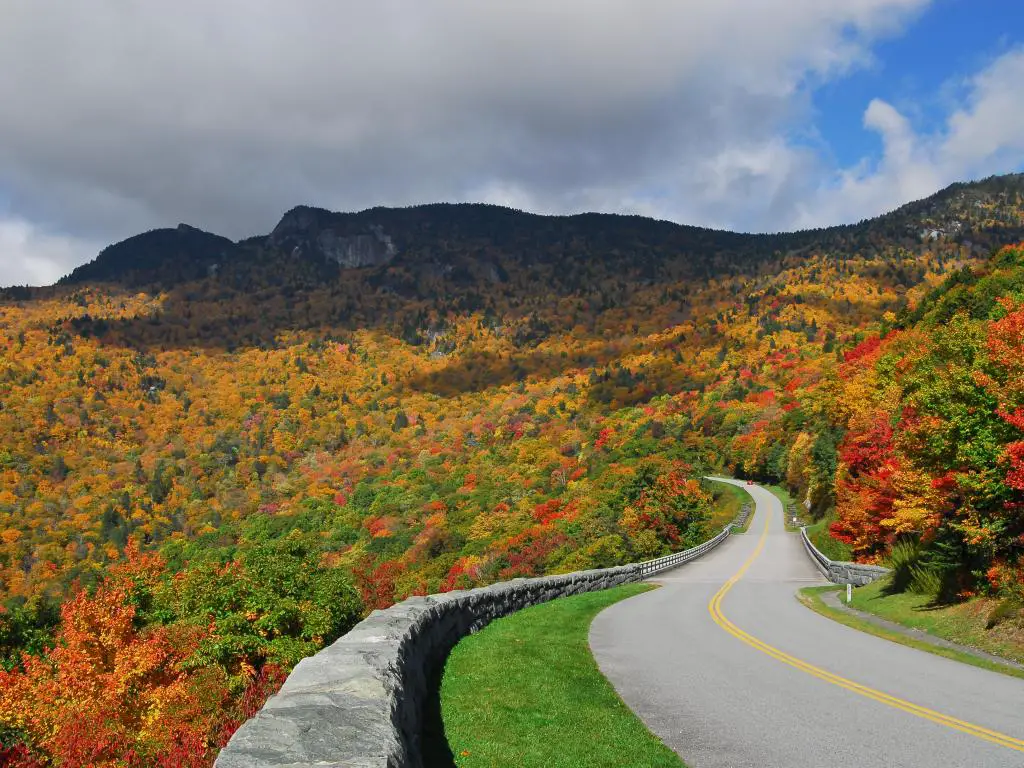 Blue Ridge Parkway, EE.UU. con vistas a las montañas Blue Ridge a lo lejos bajo un cielo nublado pero azul, con la carretera que conduce hacia los árboles en otoño.
