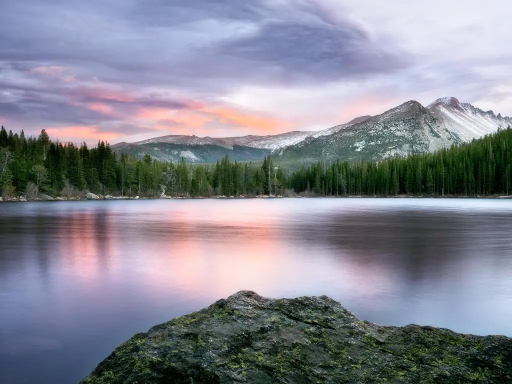 Agua gris plana en un amplio lago que refleja la luz rosa del atardecer con montañas cubiertas de nieve detrás y pinos en las orillas del lago