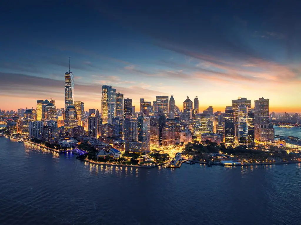 El horizonte panorámico de la ciudad de Nueva York al amanecer con los edificios iluminados en amarillos y naranjas y el mar de color azul oscuro. 