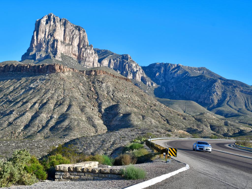 Pico rocoso se eleva por encima de la carretera con un coche, con un cielo azul claro detrás