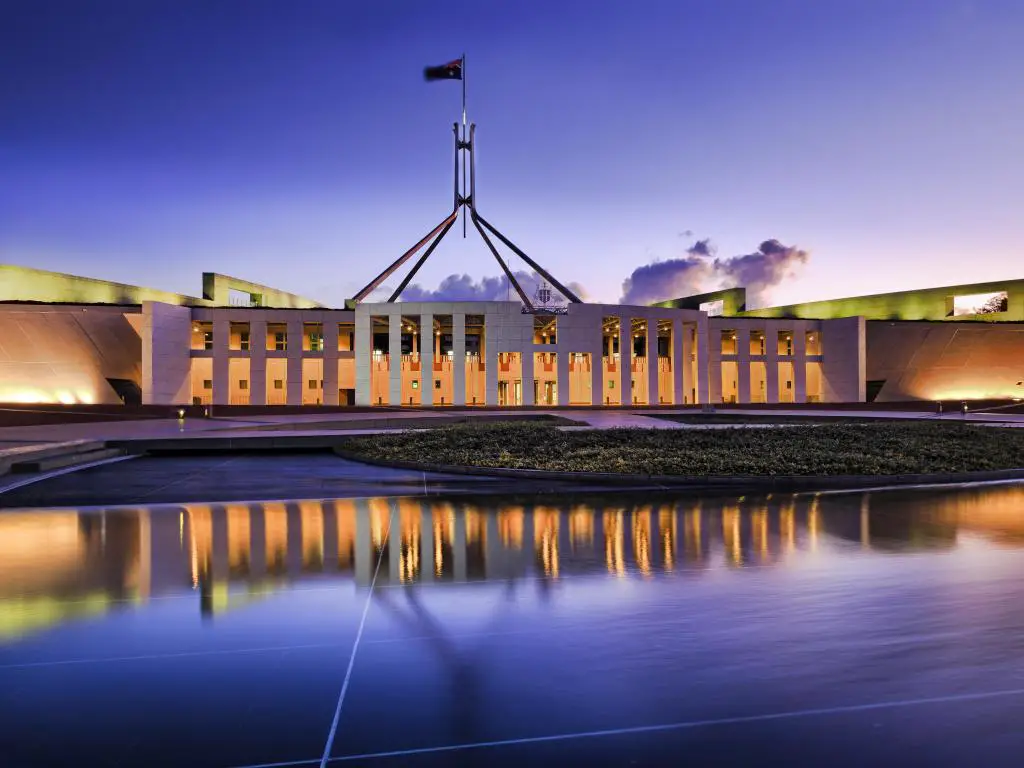 Fachada de la Casa del Parlamento Nacional Australiano, Canberra, brillantemente iluminada y reflejada en el agua borrosa del estanque de la fuente bajo la bandera nacional ondeando en el asta de la bandera al atardecer.