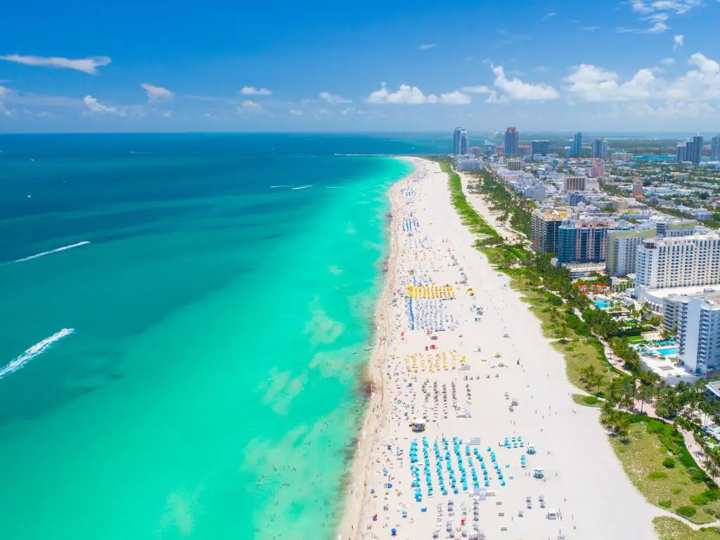 Vista aérea de Miami Beach, Florida, del largo tramo de playa con tumbonas, a ambos lados de un mar turquesa y los edificios de la ciudad a la derecha.