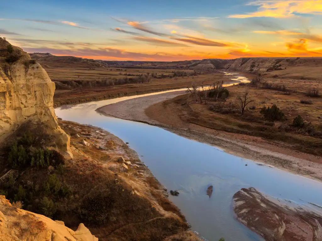 Parque Nacional Theodore Roosevelt, Dakota del Norte con el río Little Missouri atravesando, formaciones rocosas a un lado y llanuras planas en el otro que conducen a la distancia al atardecer con un cielo impresionante.