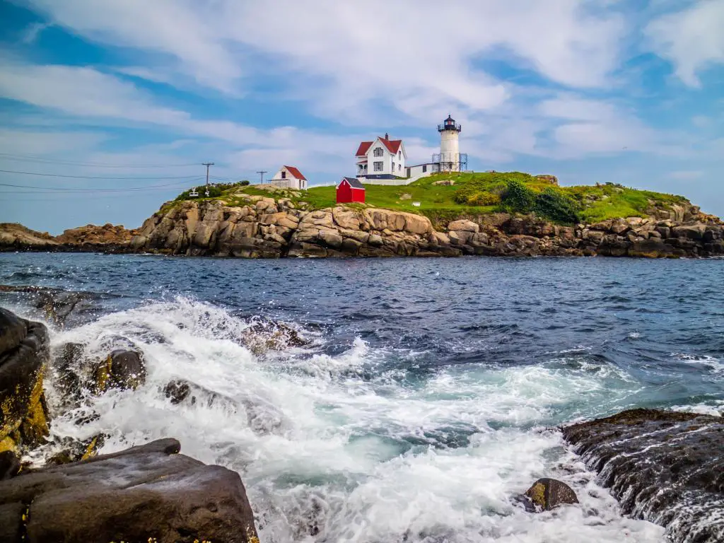 Nubble Lighthouse, Maine USA con un mar golpeando rocas en primer plano y una isla en el horizonte con un faro y algunos edificios salpicados a su lado en un día nublado.