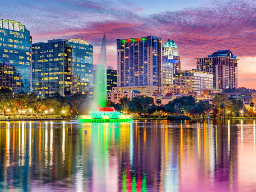El chorro de la fuente iluminado en verde brota del agua del lago con el paisaje urbano y el colorido cielo de la puesta de sol de lana de algodón