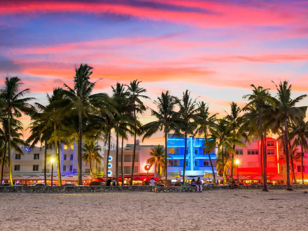 Miami Beach, Florida, en Ocean Drive al atardecer, con una playa en primer plano y palmeras ante una hilera de edificios iluminados con colores fluorescentes.