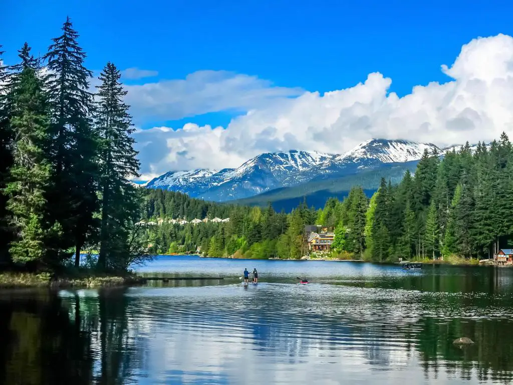 Whistler, Columbia Británica, Canadá, con vistas al lago Alta en primer plano y árboles que bordean la orilla con montañas cubiertas de nieve en la distancia en un día soleado.