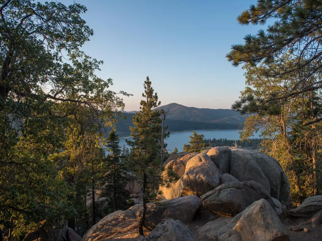 Big Bear Lake, California, EE.UU. con grandes rocas en primer plano y árboles y el lago y las montañas más allá tomadas al amanecer.