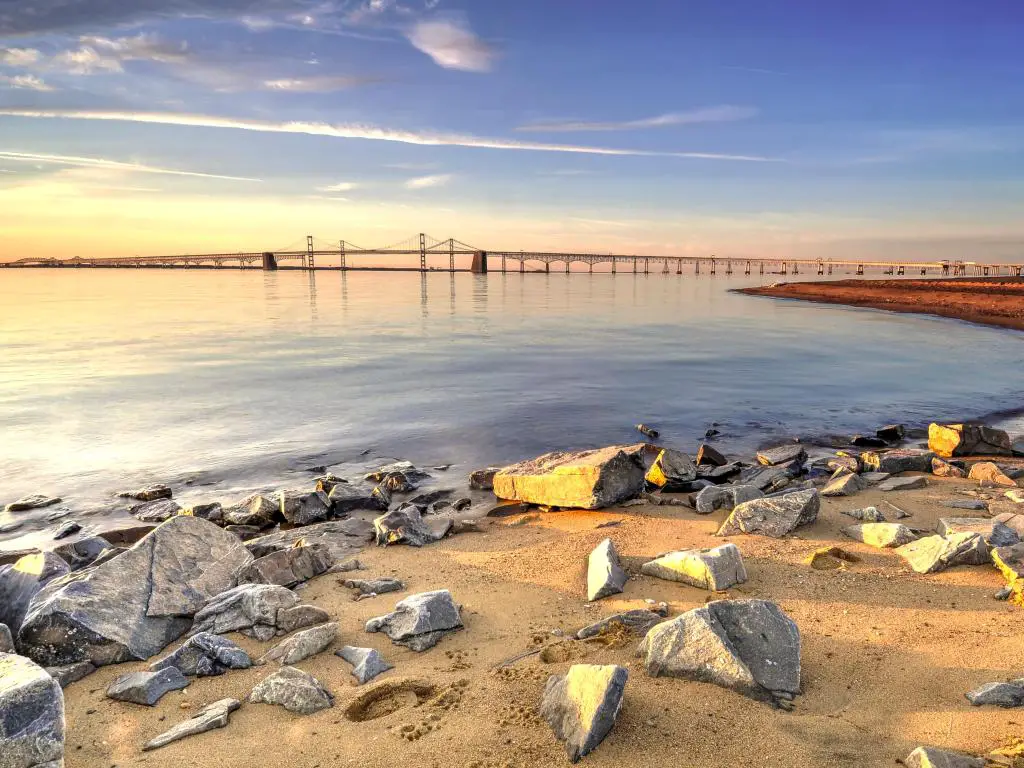 Puente de la Bahía de Chesapeake, Bahía de Chesapeake, EE. UU. Tomado al amanecer con un colorido cielo matutino sobre el Puente de la Bahía de Chesapeake y la playa en primer plano con una variedad de piedras yacen pacíficamente, sumergidas en la luz del sol dorada.