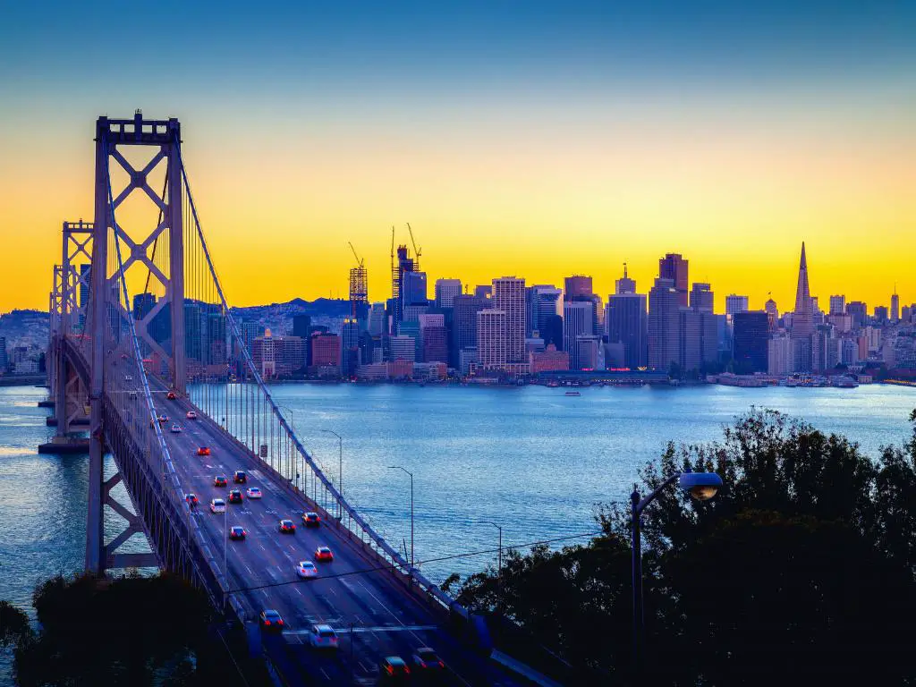 Un hermoso resplandor crepuscular dorado del atardecer de verano que ilumina el horizonte de San Francisco y una vista panorámica del famoso Puente de la Bahía de Oakland con autos conduciendo a lo largo del puente (Interestatal 80)