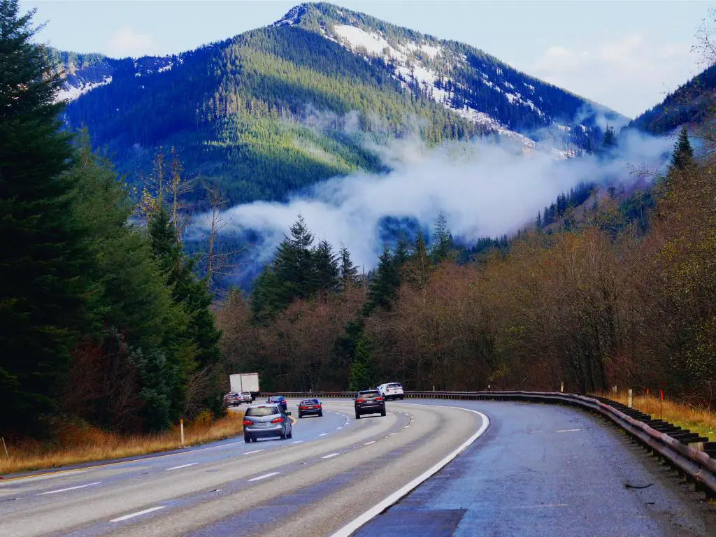 Temprano en la mañana con autos conduciendo a lo largo de Snoqualmie Pass Road pasando altos pinos verdes y disfrutando de la impresionante vista de la montaña con una delgada capa de niebla